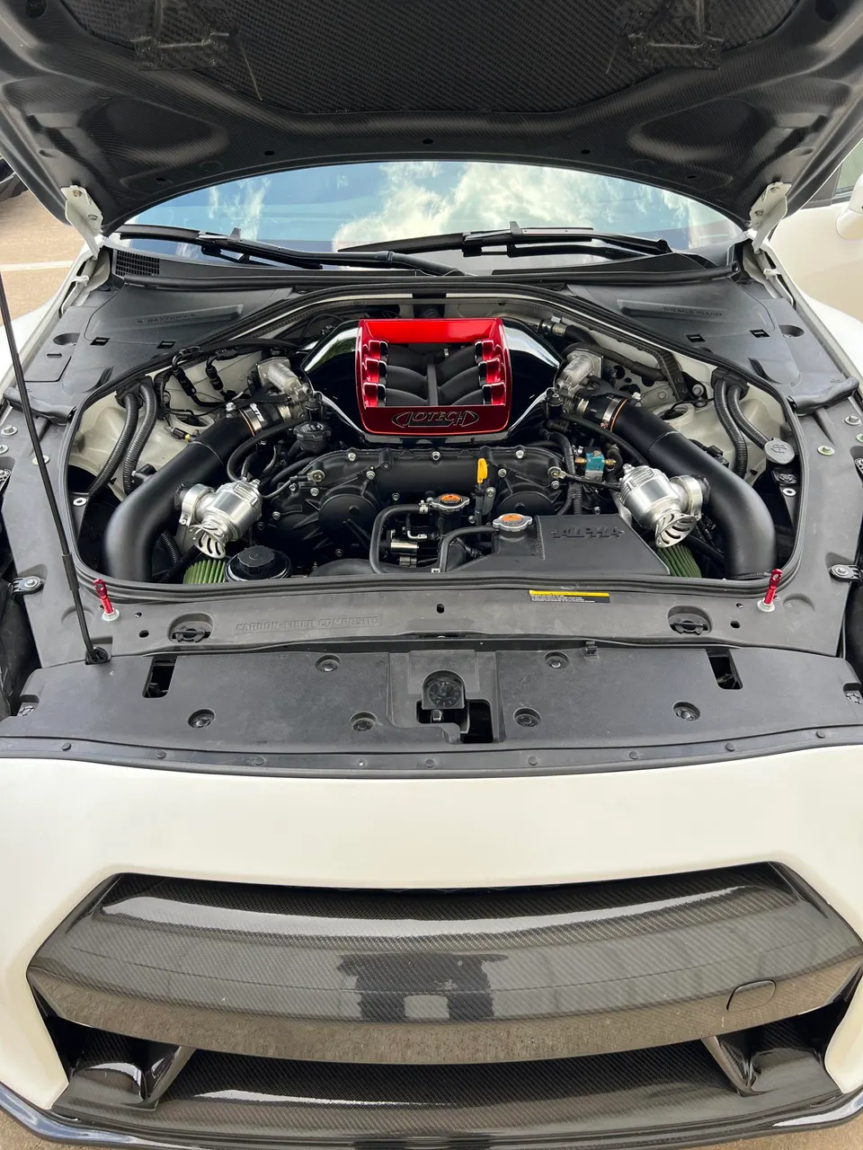 A white Nissan GT-R with its hood open, showcasing a custom red Jotech intake manifold, turbo piping, and carbon fiber hood.