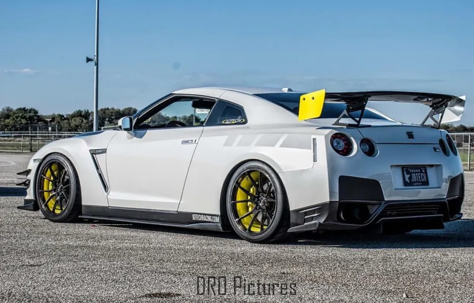 A white Nissan GT-R with a large rear wing, yellow wheels, and aero modifications parked outdoors on a sunny day.