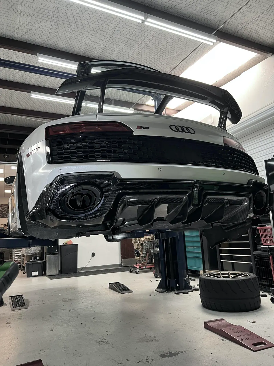 White Audi R8 on a lift in a shop, showcasing its large carbon fiber rear wing, aggressive diffuser, and exhaust.