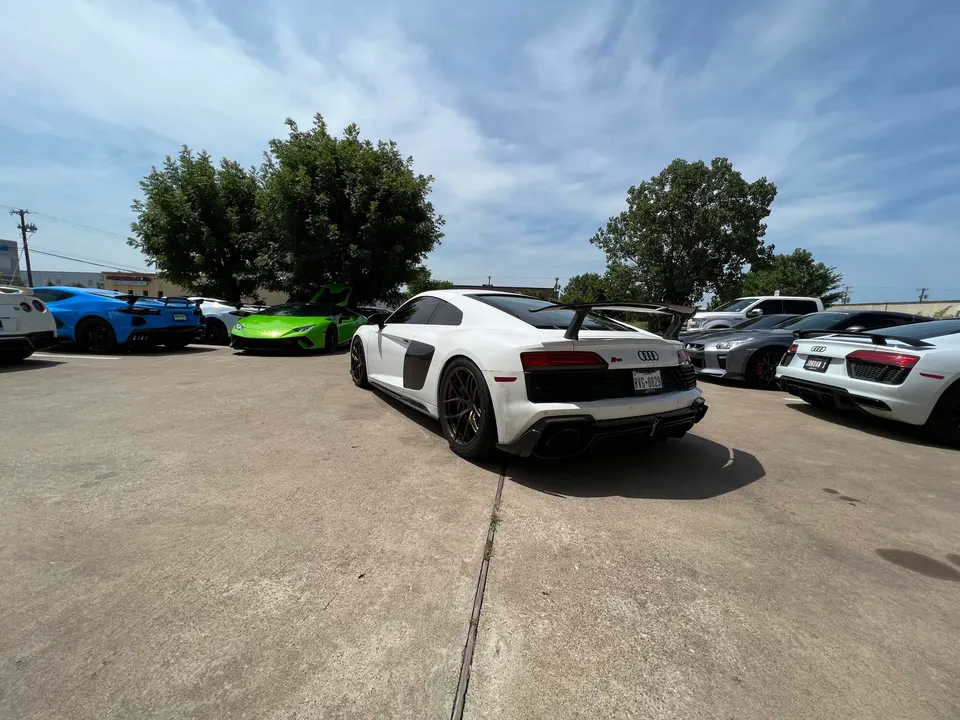 A group of high-performance cars including white Audi R8s, a green Lamborghini Huracan, a blue Corvette C8, and a grey Nissan GT-R parked outdoors.