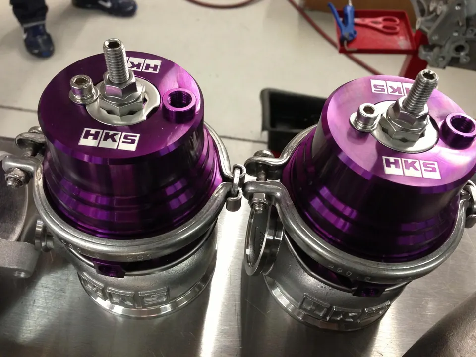 Two purple HKS wastegates on a metal workbench in a performance tuning shop, ready for installation.