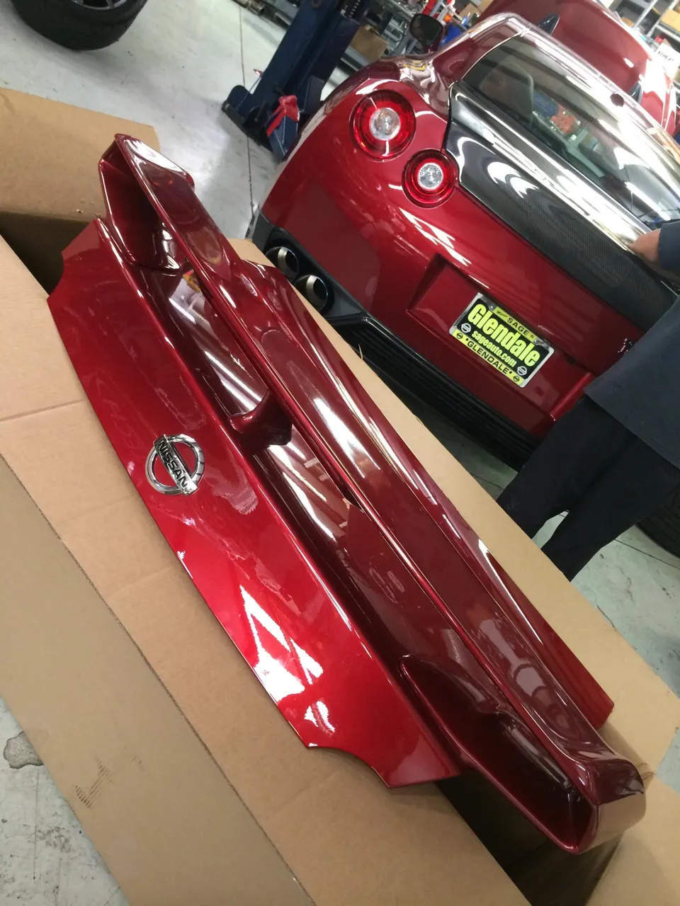 Red Nissan GT-R trunk lid with integrated spoiler in a cardboard box next to a red GT-R in a shop.