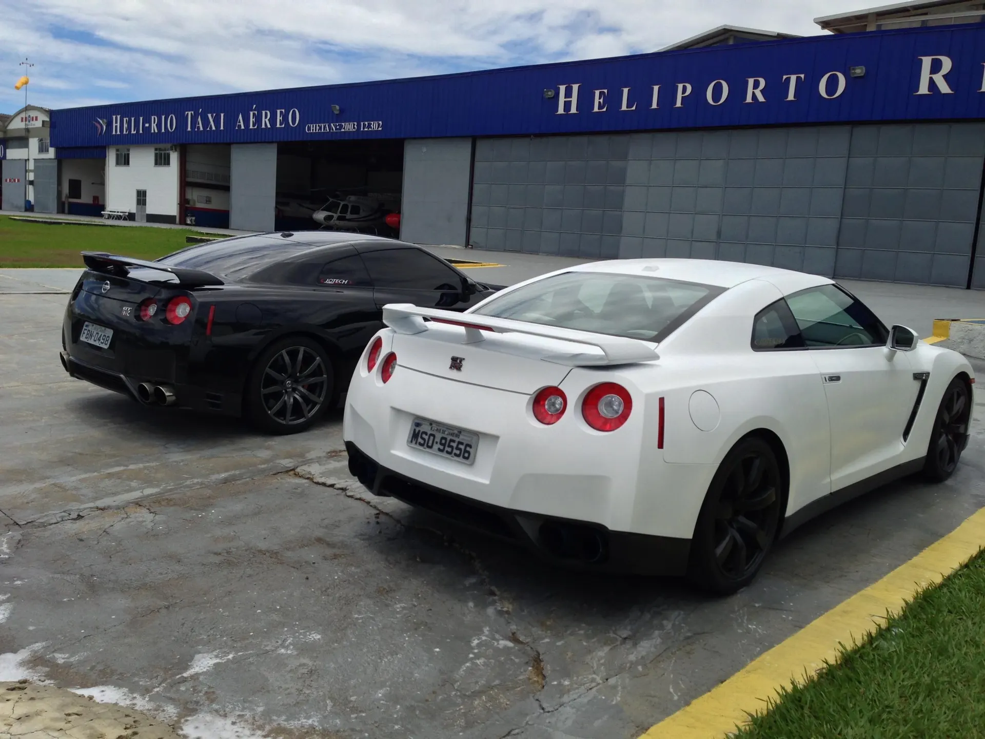 Two Nissan GT-R R35s, one black and one white, parked outside a "Heli-Rio Táxi Aéreo" hangar.