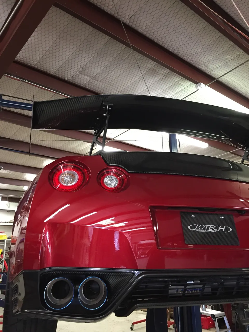 Rear view of a red Nissan GT-R on a lift in a Jotech Motorsports shop, featuring a large carbon fiber wing and custom exhaust tips.