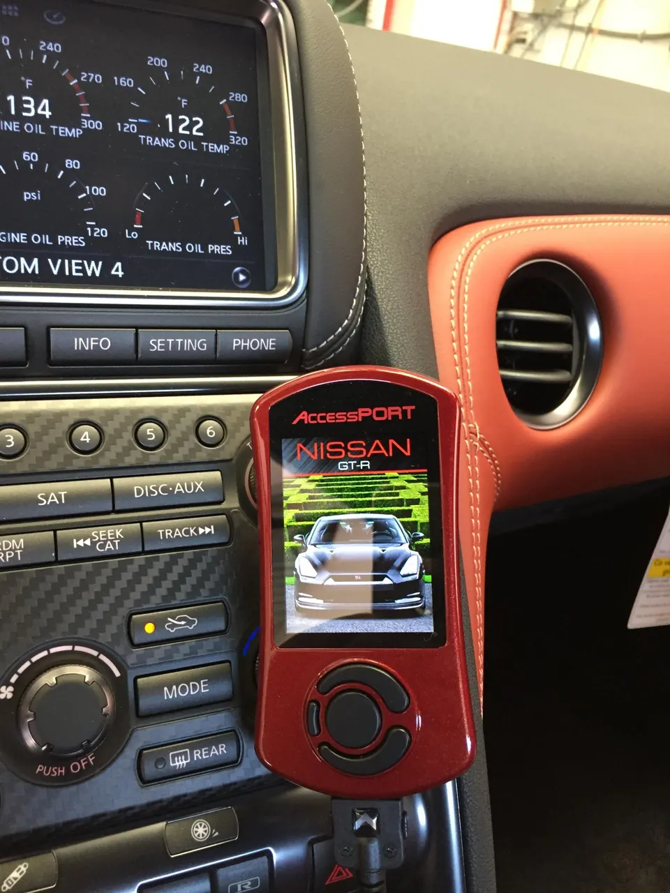 A red Cobb AccessPORT tuning device plugged into the dashboard of a Nissan GT-R, with gauges visible on the infotainment screen.