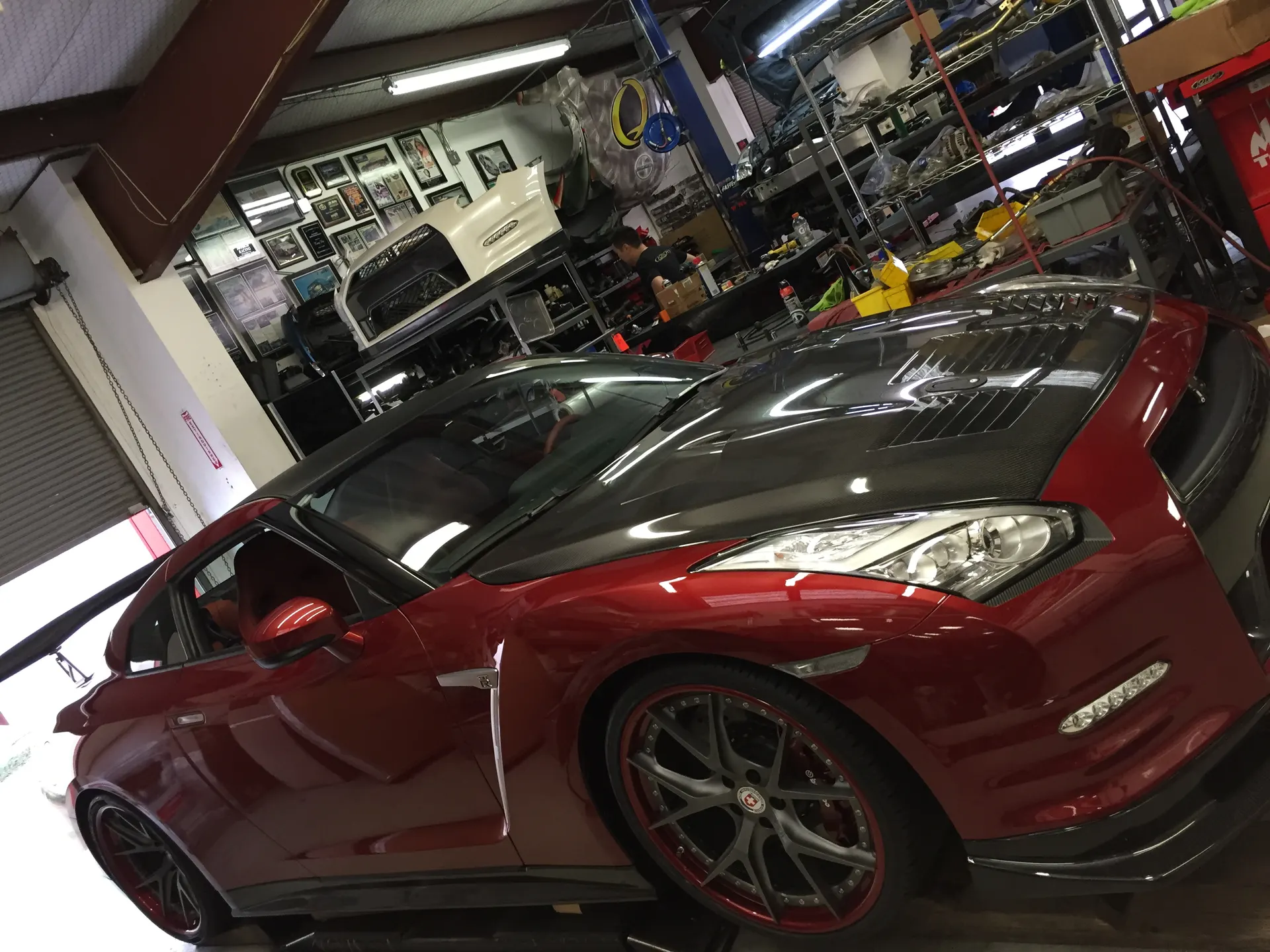Red Nissan GT-R with carbon fiber hood and aftermarket wheels inside a performance tuning shop.