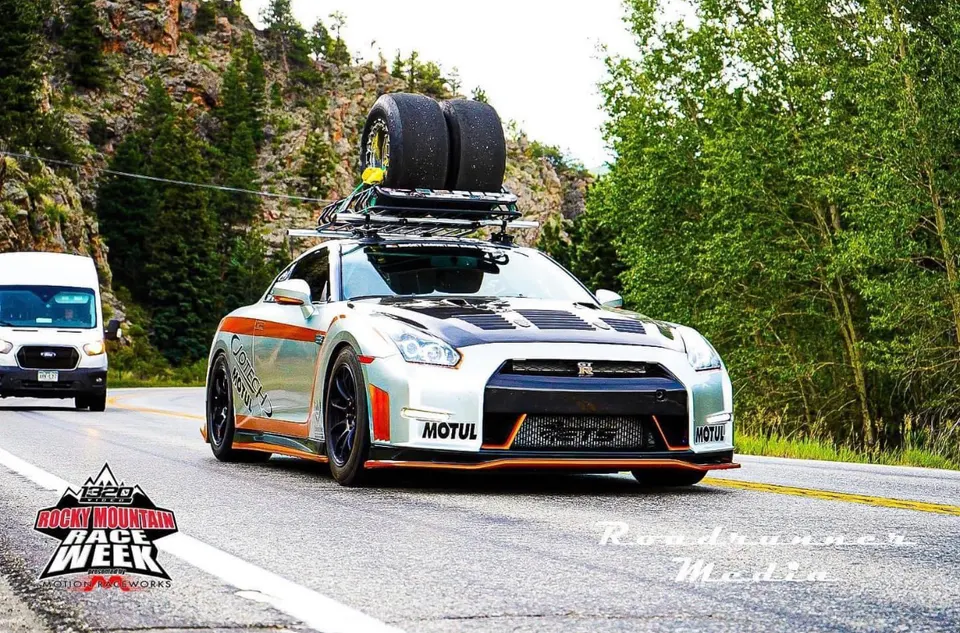 Silver Nissan GT-R with a roof rack carrying race tires drives on a scenic mountain road during Rocky Mountain Race Week.