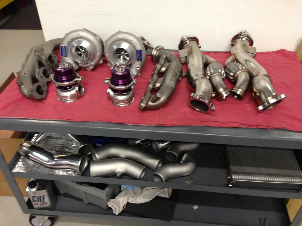 A collection of performance car parts including HKS turbochargers, purple wastegates, exhaust manifolds, and an intercooler on a shop cart.