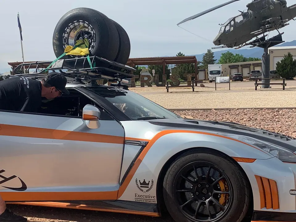 Nissan GT-R with drag tires on a roof rack, a man leaning into the car, and a helicopter in the background.