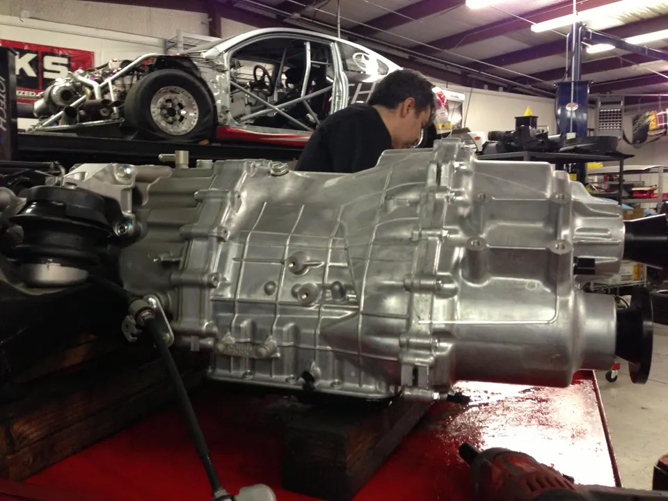 A mechanic works on a large transmission in a performance tuning shop with a race car on a lift in the background.
