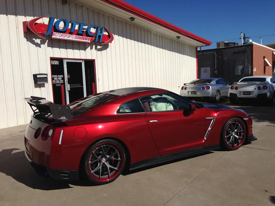 Red Nissan GT-R with carbon fiber wing and aftermarket wheels parked outside Jotech Motorsports performance shop.