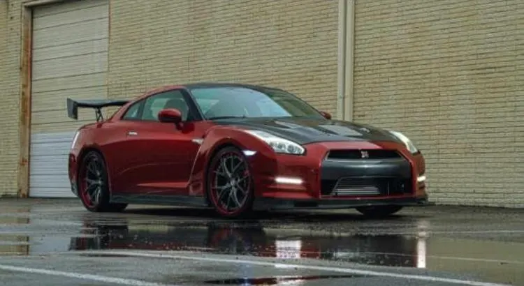 A red Nissan GT-R with a carbon fiber hood, aftermarket wheels, and a large rear wing parked on wet pavement.