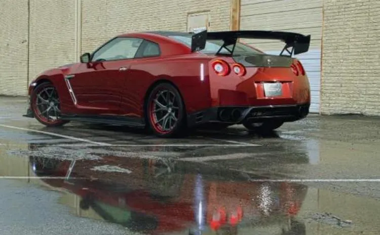 A red Nissan GT-R with a large carbon fiber wing and aftermarket wheels parked on wet pavement, reflecting in puddles.