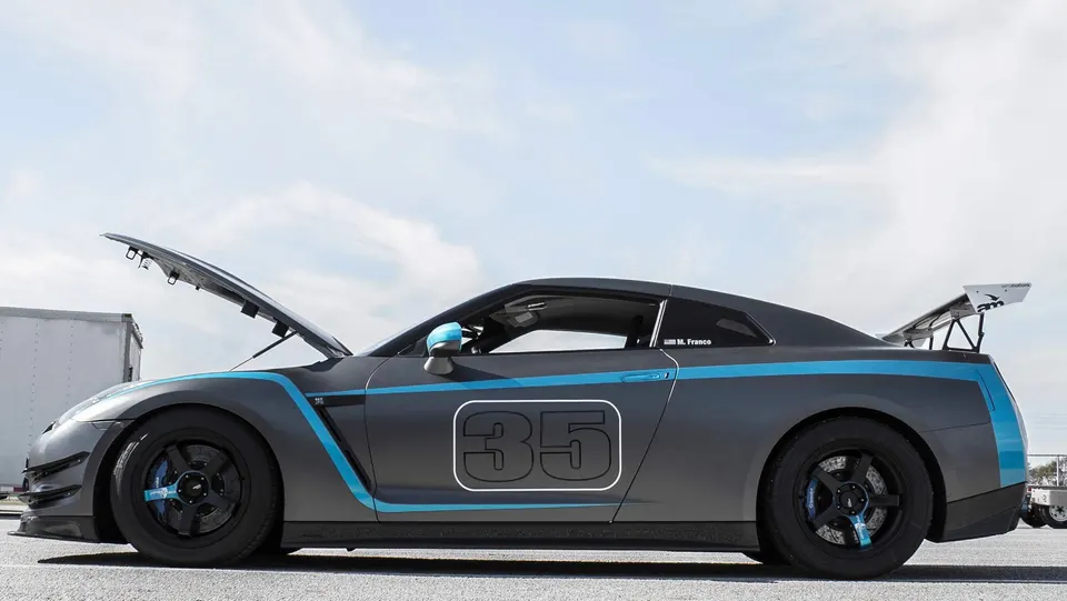 Grey Nissan GT-R R35 race car with blue accents and number 35, featuring aftermarket wheels and a large rear wing, parked at a track with its hood open.