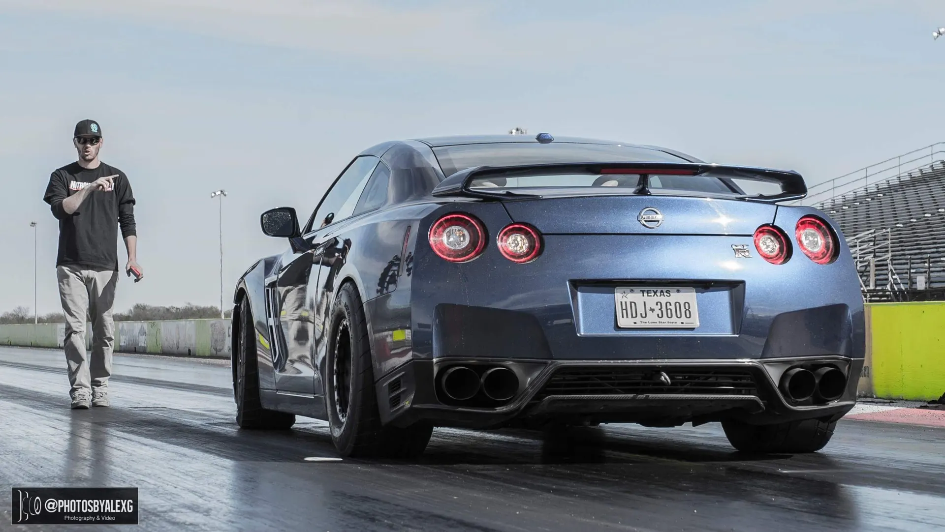 A blue Nissan GT-R with drag tires on a drag strip, with a person walking beside it.