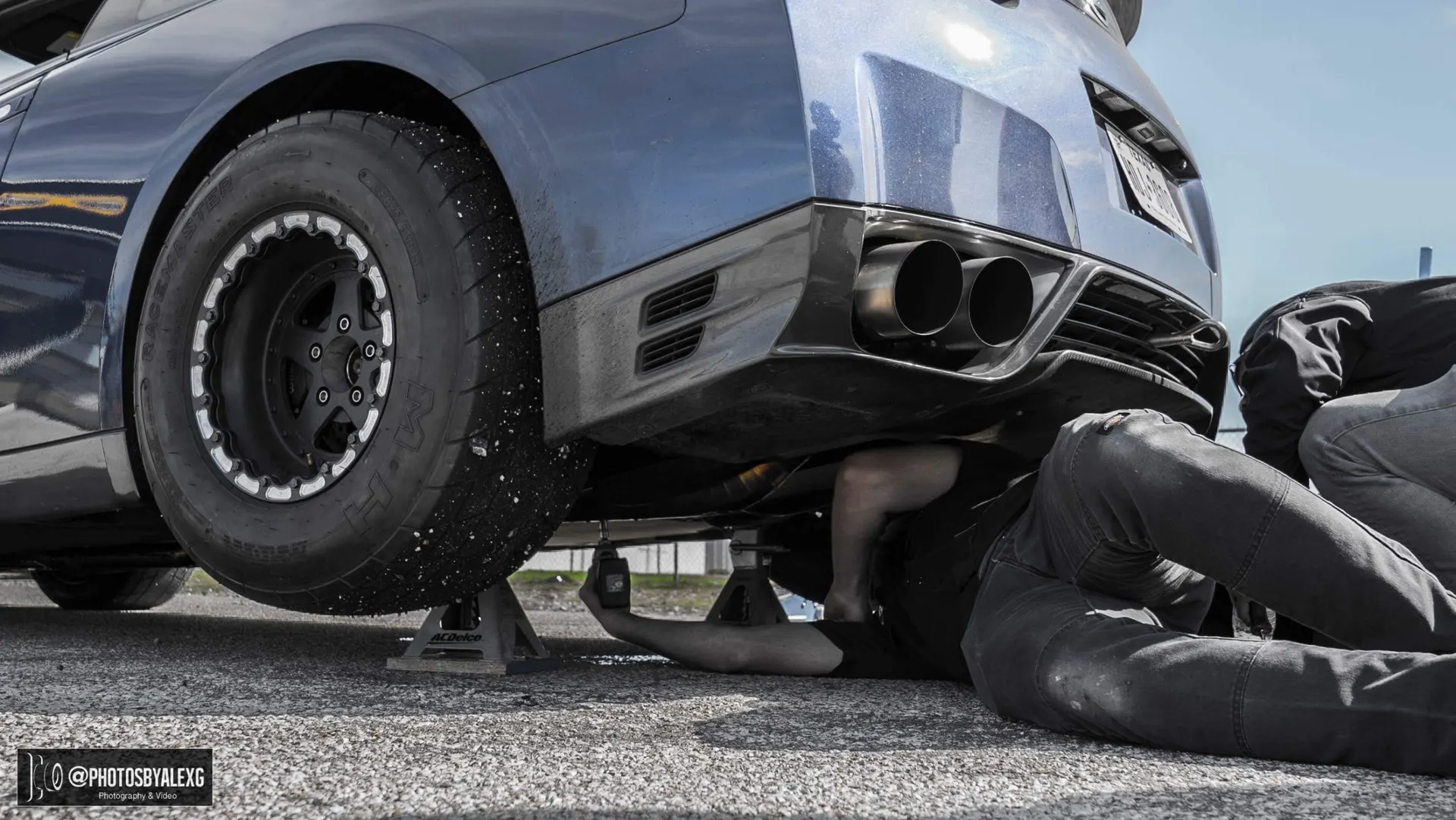 Nissan GT-R on jack stands with a mechanic working underneath, featuring a drag tire and aftermarket exhaust system.