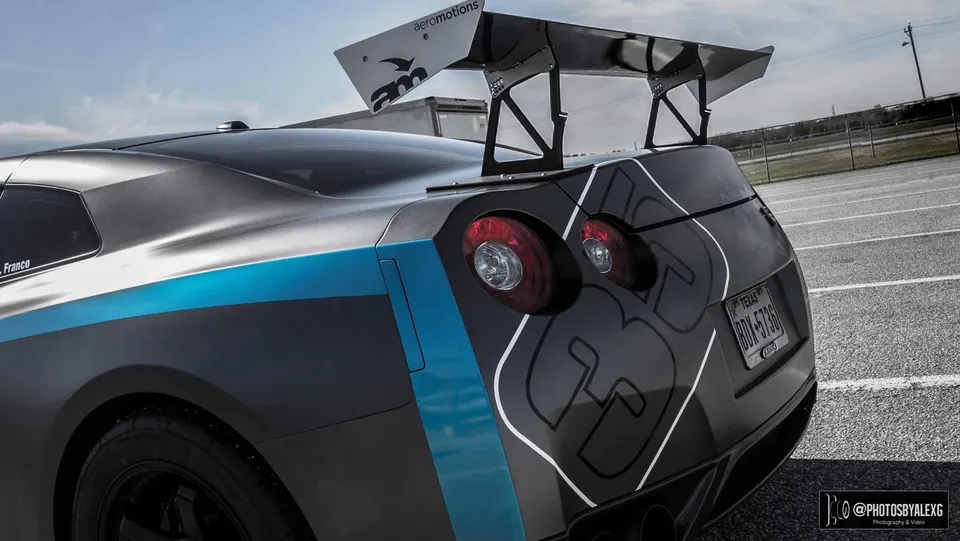 A matte grey Nissan GT-R with a blue stripe and a large Aeromotions rear wing parked outdoors.