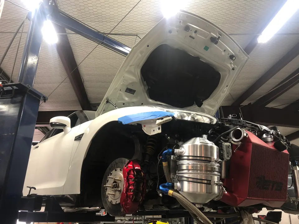 A white Nissan GT-R on a lift in a shop with its hood open, showcasing a large red intercooler and custom engine components.