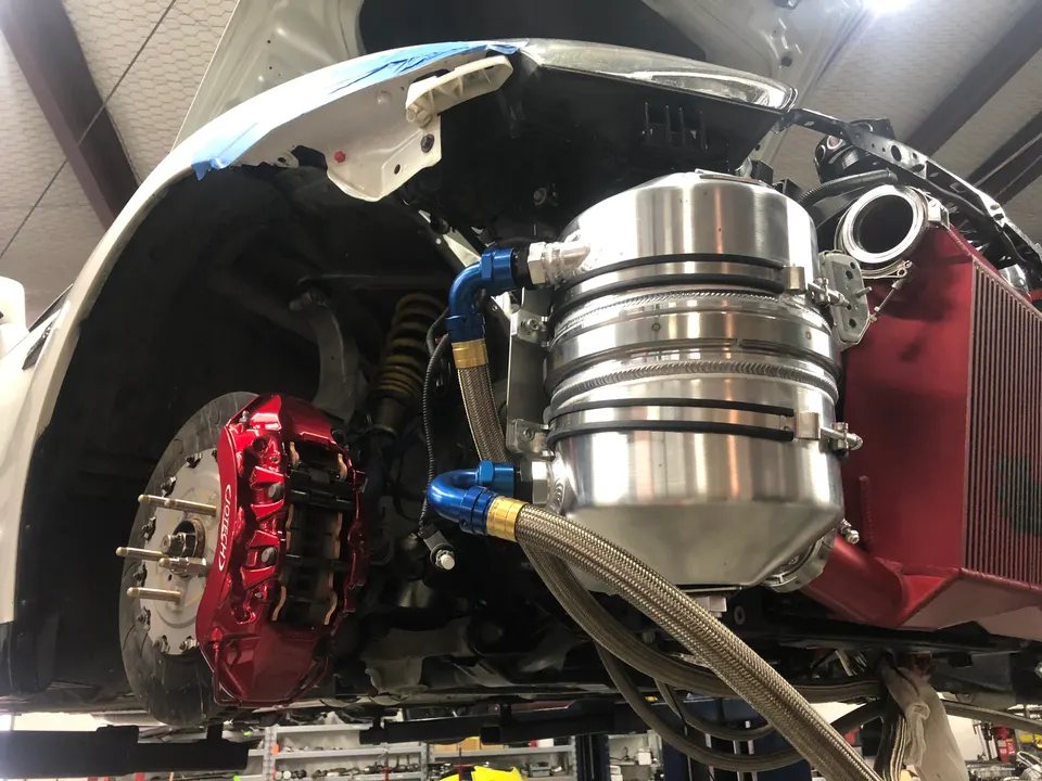 Front end of a performance car on a lift, showcasing a large intercooler, custom cooling system with braided lines, and red performance brakes.