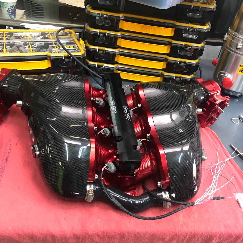 A carbon fiber intake manifold with red anodized components and Alpha Performance fuel rails on a workbench in a shop.