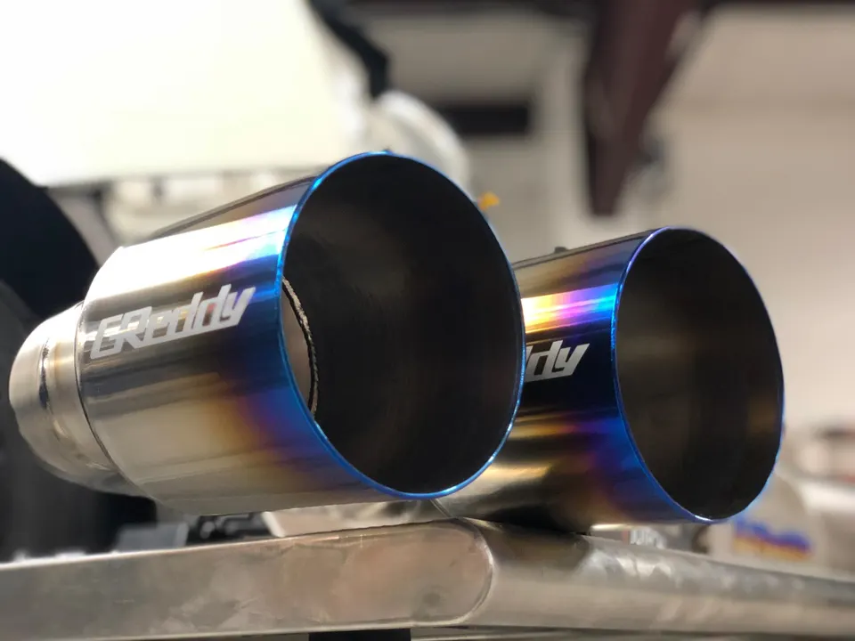 Close-up of GReddy titanium exhaust tips with a burnt finish, showcasing aftermarket performance parts.
