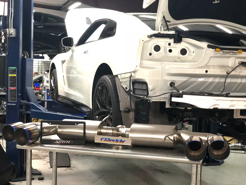 White Nissan GT-R on a lift in a shop with its rear bumper removed, next to a GReddy Jotech exhaust system.
