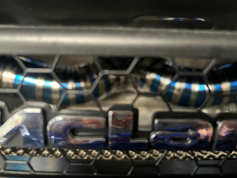 Close-up of blue heat-treated titanium exhaust piping visible through a car's front grille with a partial badge.