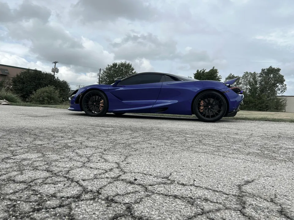 Blue McLaren 720S supercar parked outdoors on an asphalt lot under a cloudy sky, showcasing its sleek side profile.