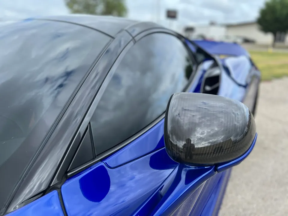 A close-up of a blue supercar's carbon fiber side mirror, reflecting the sky and surroundings.