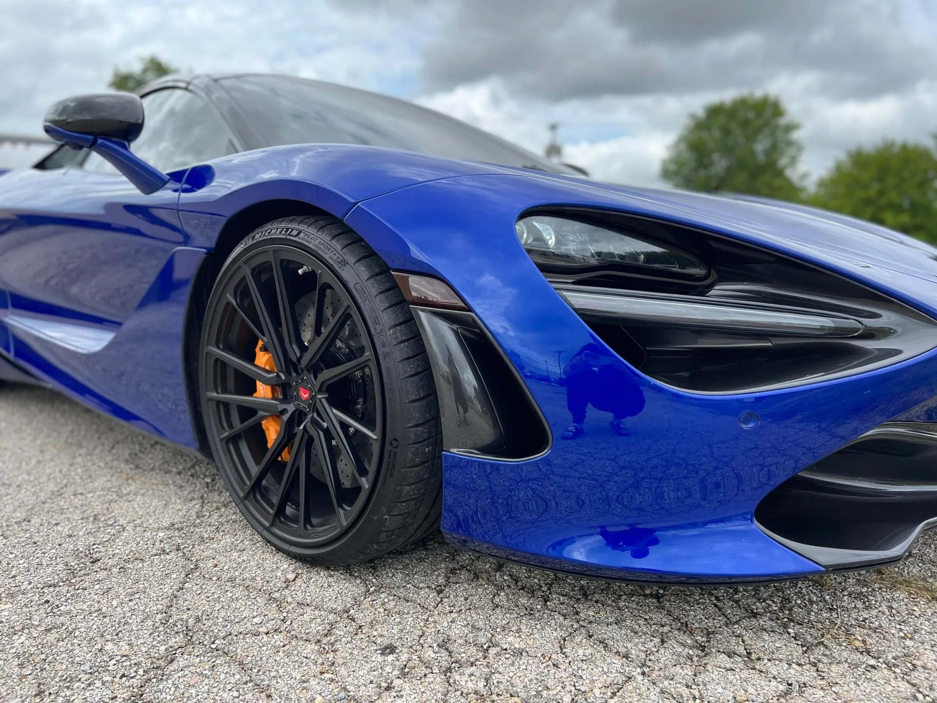 A blue McLaren 720S with black aftermarket wheels and orange brake calipers, parked outdoors.