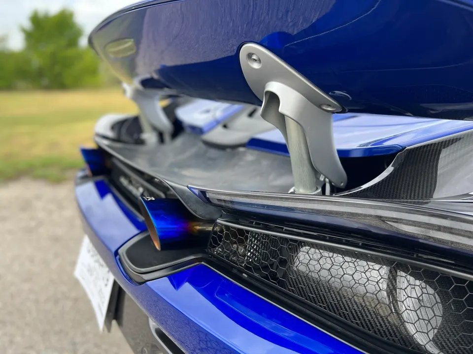 A close-up of the rear of a blue McLaren supercar, showcasing its active aerodynamic wing, carbon fiber details, and a vibrant blue exhaust tip.