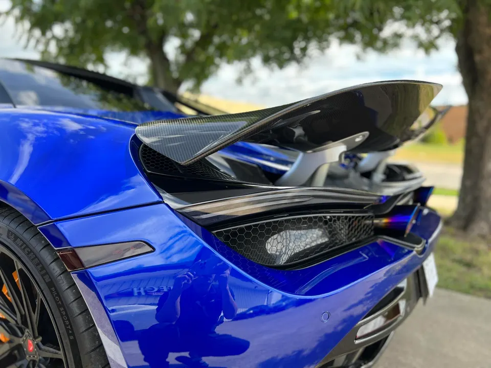 Rear view of a blue McLaren supercar, showcasing its carbon fiber active aero wing and titanium exhaust tips.