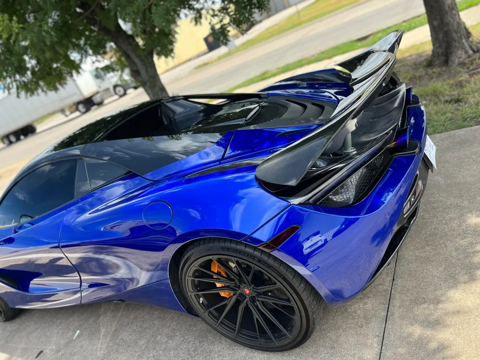 A blue McLaren 720S with a large carbon fiber rear wing and aftermarket wheels parked outdoors.