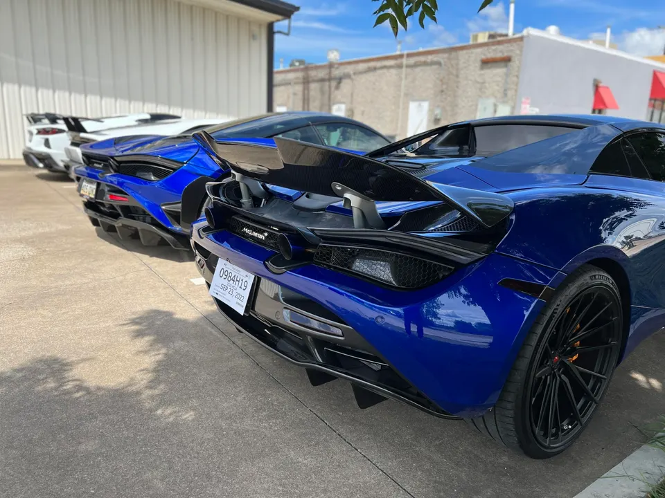 Rear view of multiple McLarens, including a blue McLaren 720S with a carbon fiber wing, parked outdoors.