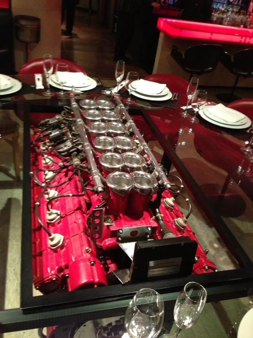 A red V12 engine with chrome velocity stacks displayed as a glass-top dining table in a restaurant setting.