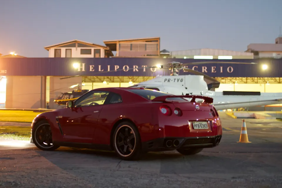 Red Nissan GT-R parked at a helipad with a helicopter in the background at dusk.