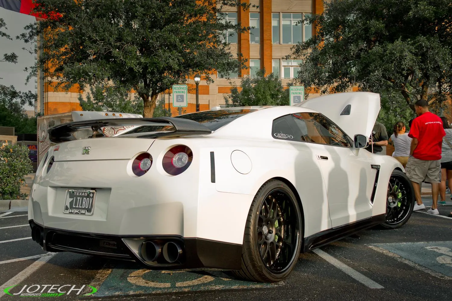 White Nissan GT-R with an open hood and aftermarket wheels parked at an outdoor car event with people around.