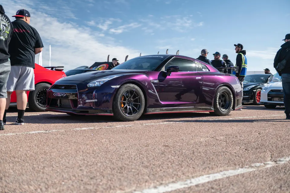 A vibrant purple Nissan GT-R (R35) with aftermarket wheels and aero, parked at a track day event with people around.