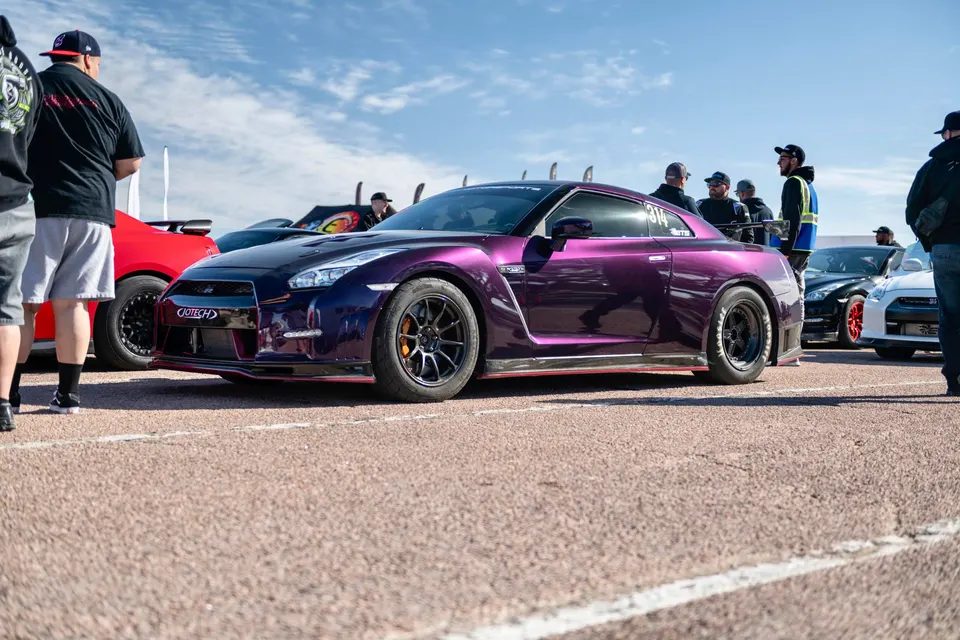 A purple Nissan GT-R with aftermarket wheels and aero parked at a track event with people around under a blue sky.