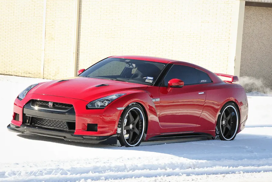 Red Nissan GT-R R35 with aftermarket wheels and front splitter parked in the snow against a brick wall.