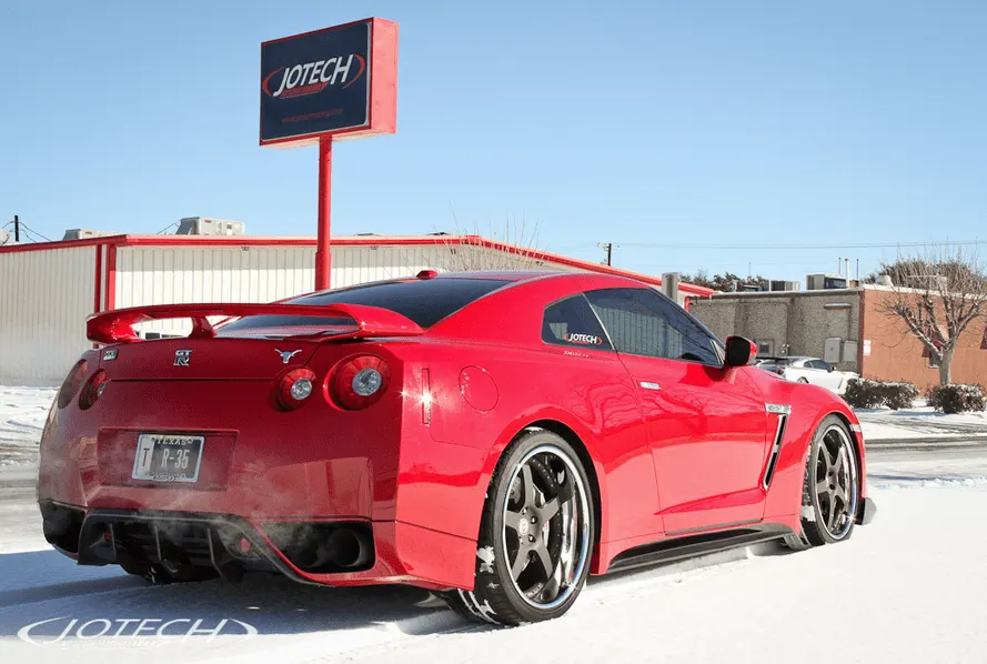 Red Nissan GT-R with aftermarket wheels and rear wing parked in snow outside Jotech Motorsports shop.