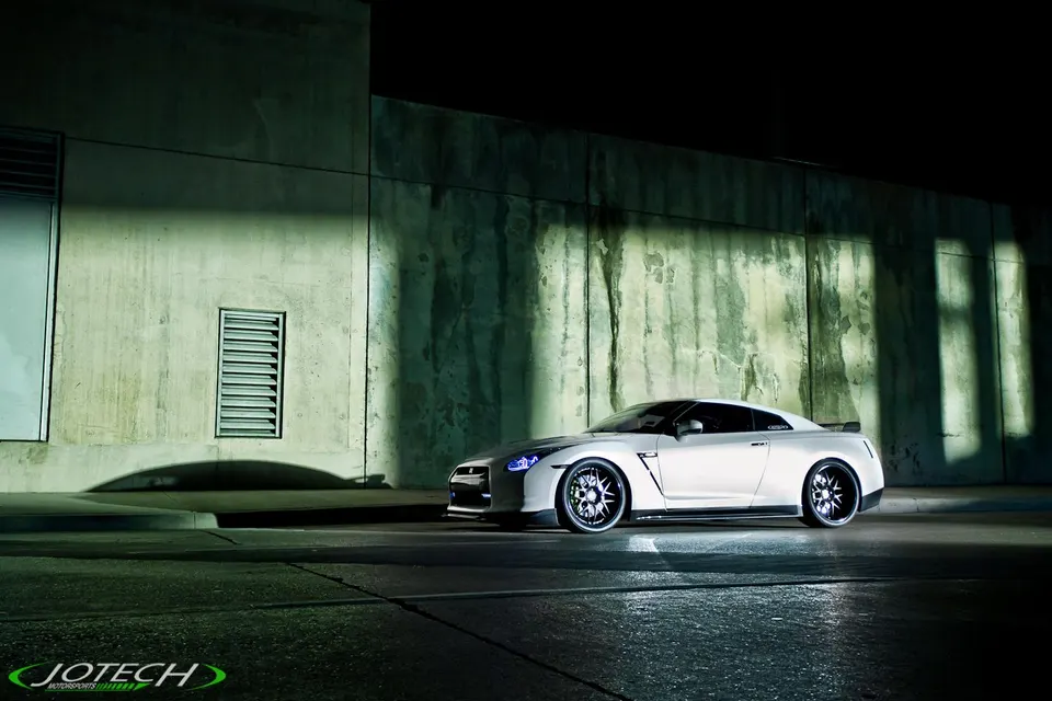 White Nissan GT-R with custom wheels parked at night against a concrete wall, dramatically lit.