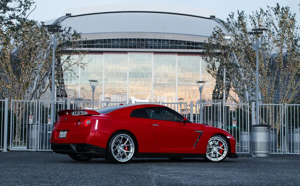 A red Nissan GT-R sports car with aftermarket wheels parked in front of a modern stadium building with trees lit with lights.