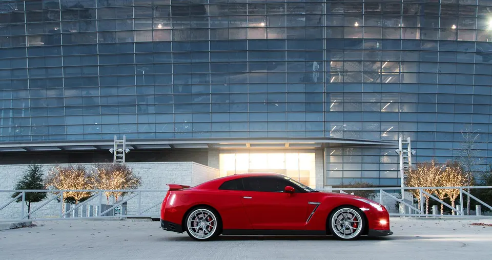 Red Nissan GT-R sports car with aftermarket wheels parked in front of a modern glass building.