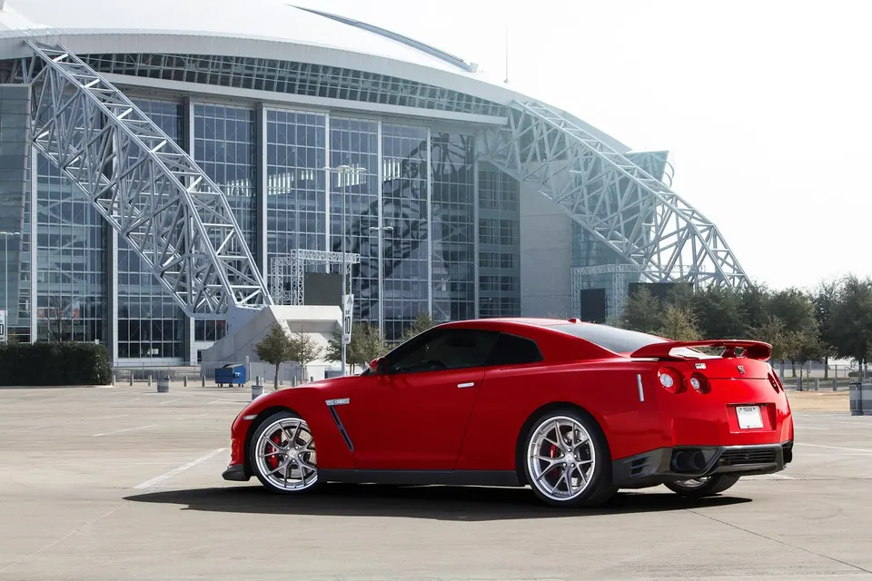 Red Nissan GT-R with aftermarket wheels parked in front of a modern stadium building.