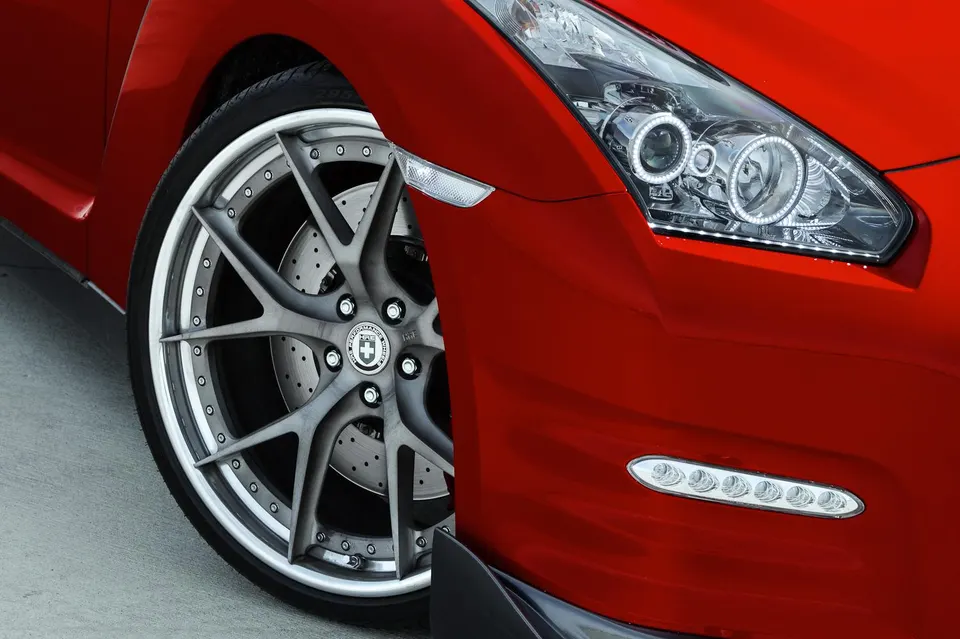 A close-up of a red Nissan GT-R's front wheel, custom HRE performance wheel, drilled brake rotor, and LED headlights.