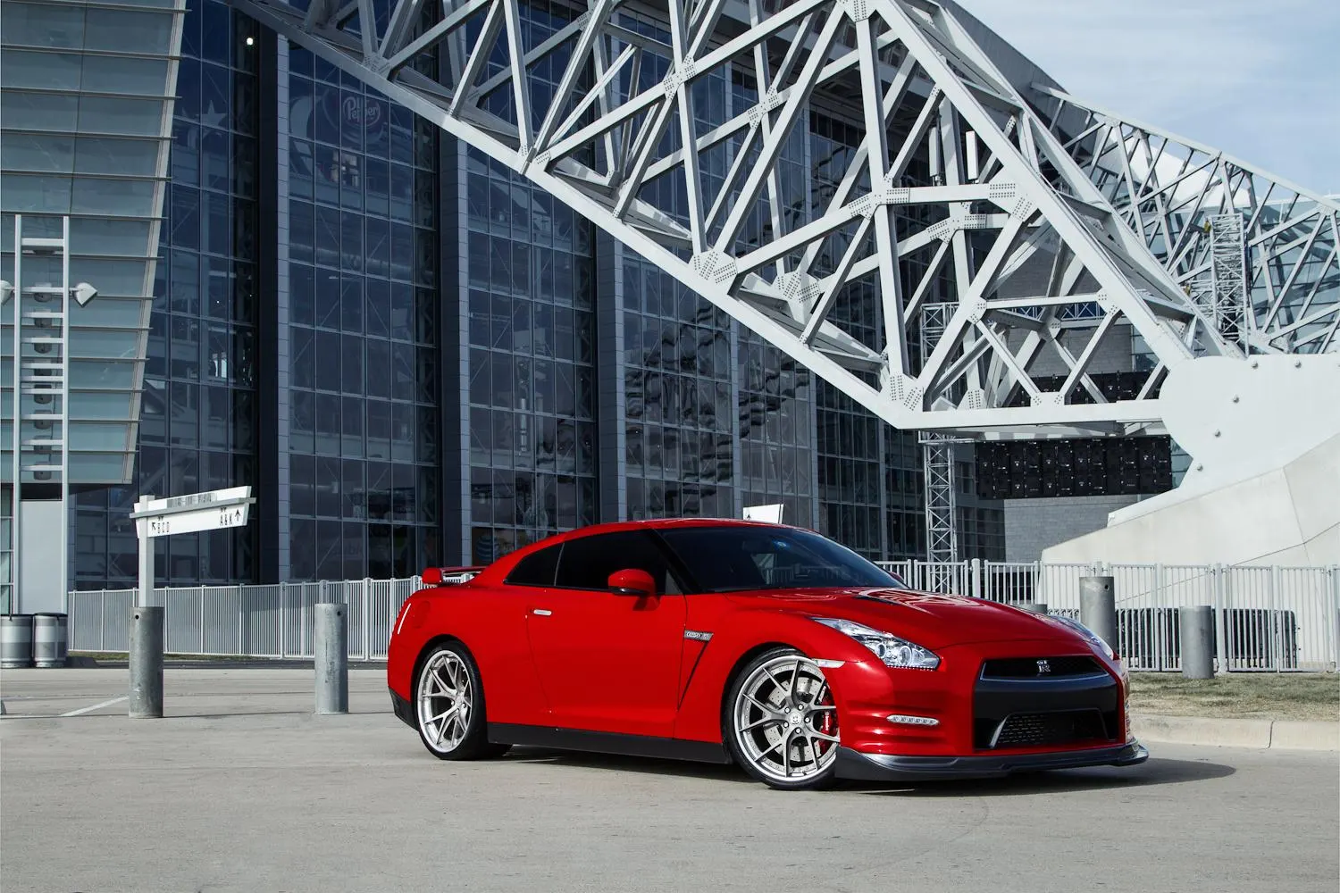 A red Nissan GT-R with aftermarket wheels parked in front of a modern glass building with a large metal structure.