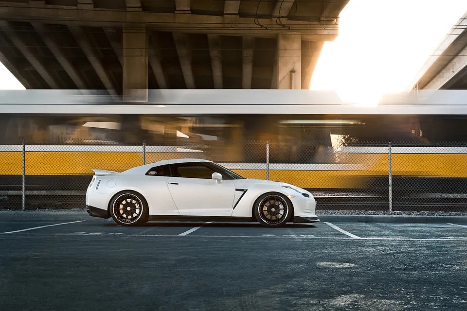 White Nissan GT-R with aftermarket wheels parked in an urban setting with a train passing by under an overpass.
