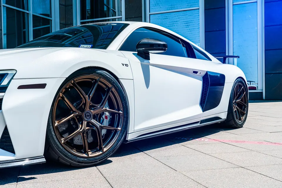 A white Audi R8 with bronze aftermarket wheels parked in front of a modern building with blue reflective windows.