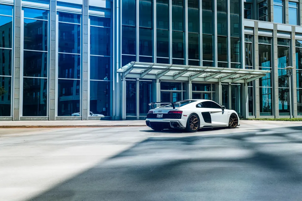 White Audi R8 with an aftermarket rear wing and bronze wheels parked in front of a modern glass building.